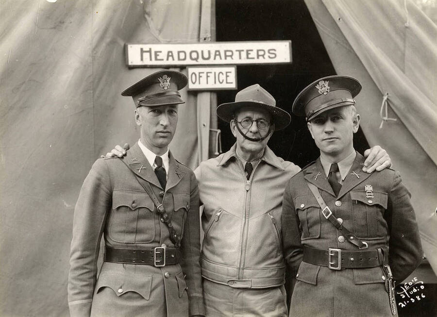 Group portrait of CCC men in front of the Headquarters Office tent at Hayden Creek CCC Camp. Sign reads: 'Headquarters Office'. Writing on the photo reads: Leo's Studio'.