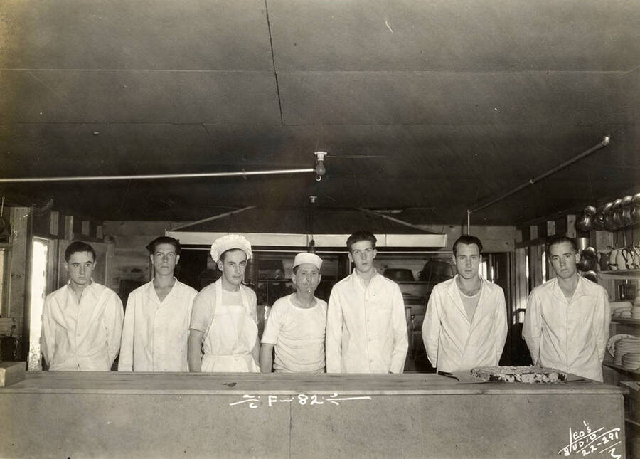 Group portrait of CCC men in the kitchen of Wolf Lodge Bay CCC Camp. Writing on the photo reads: 'F-82 Leo's Studio'. Back of photo reads: 'Wolf Lodge Beauty Bay Richardson'.