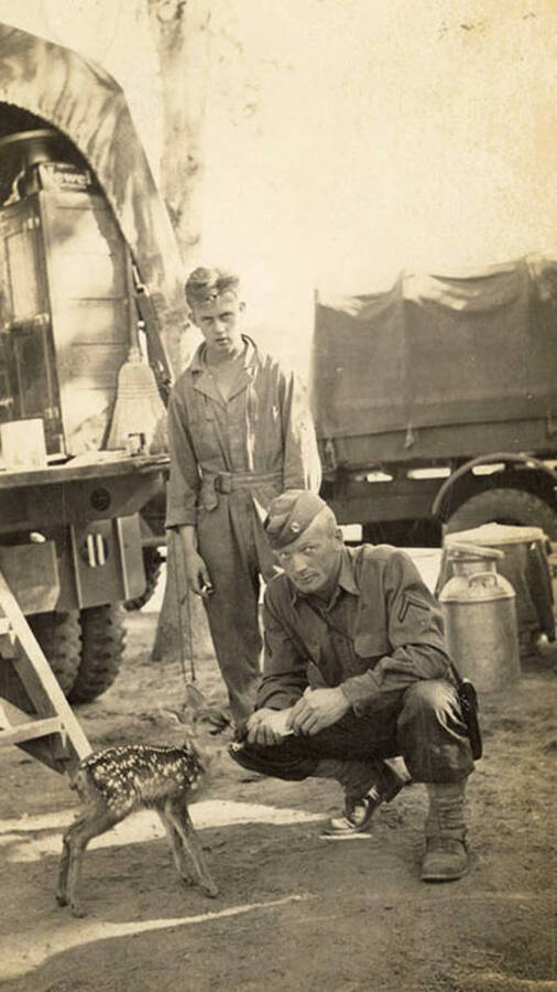 Group portrait of two CCC men at the Wolf Lodge Bay CCC Camp. The two men are posed with a pet fawn. Back of photo reads: 'Beauty Bay left to right Ralph McGraw behind Bill McGinnis truck driver.'