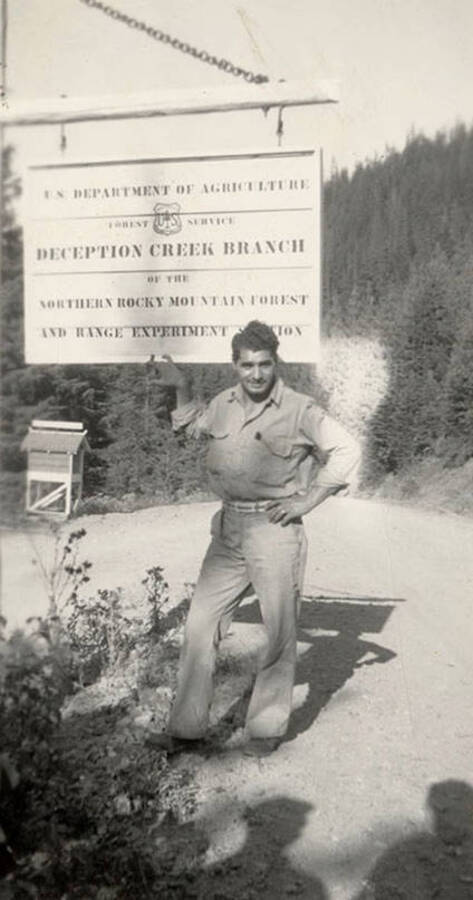 Portrait of a CCC man posing with a sign that reads: 'U.S. Department of Agriculture Forest Service Deception Creek Branch of the Northern Rocky Mountain Forest and Range Experiment Station'. Man has been identified as Salvature R. Triassi.