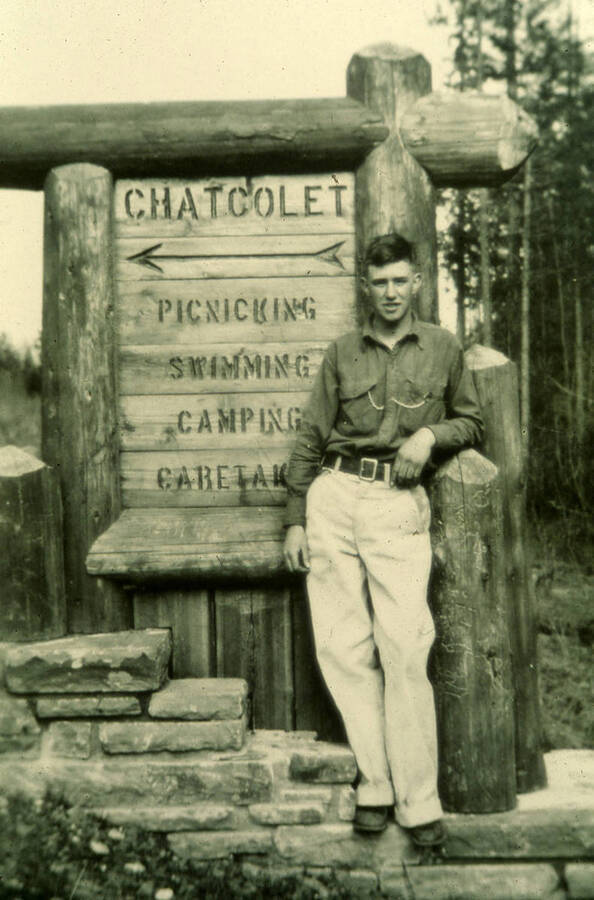 A man stands in front of a sign advertising for Lake Chatcolet. The sign reads 'Chatcolet Picnicking Swimming Camping Caretak.' The man is standing in front of the last word which may be assumed to be 'Caretaking.'