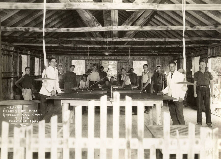 Group portrait of CCC men playing pool and ping pong in the recreation hall at Grizzly CCC Camp, F-136. Writing on the photo reads: 'Recreation Hall Grizzly CCC Camp F-136 Enaville, Idaho. Leo's Studio'.