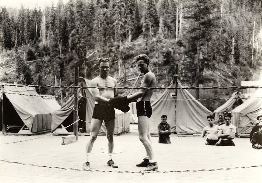 Two CCC men posed for a photo in a boxing ring while other men look on at the Deception Creek CCC Camp, F-137. Writing on photo reads: 'Leo's Studio'. Back of photo reads: 'Left A. Kelley H. Rose Deception F-137 Company 545'.