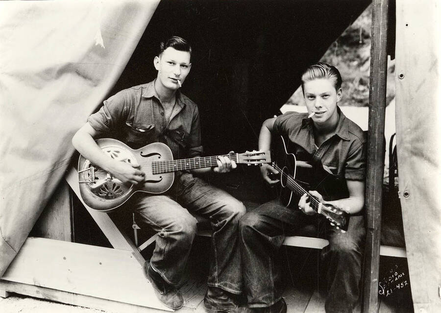 Two CCC men playing guitars and smoking cigarettes in a tent barrack at the Deception Creek CCC Camp, F-137. Writing on the photo reads: 'Leo's Studio, Spokane'. Back of photo reads: 'Max Waugh and Ralph Loyd Deception F-137 Company 545'.