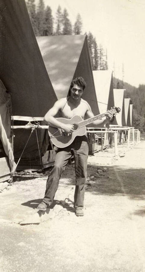 Salvature R. Triassi playing the guitar in front of tent barracks at the Deception Creek CCC Camp, F-137. Writing on the photo reads: 'Deception Creek'.