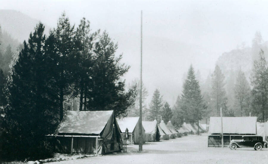 View of tent camp surrounded by trees. Flagpole in the center and an automobile is parked by one of the tents.