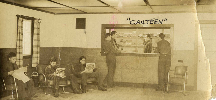 Group of CCC men standing around and reading newspapers at the Canteen at CCC Camp Big Creek #2. The canteen was at one end of the Recreation Room which had ping pong tables and was where most of the men hung out in their spare time. Norman Perkins is behind the counter. Writing on the photo reads ''Canteen' CCC Company 531 Camp Big Creek #2 F-132 Prichard, ID'.