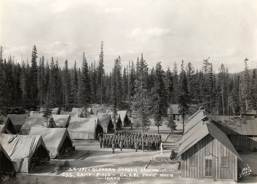 View of Gleason Ranger Station CCC Camp, F-162 and the CCC men standing at attention. The sign over the door on the building on the right hand side of the photo reads: 'headquarters'. Writing on the photo reads: 'Gleason Ranger Station CCC Camp F-162 Company 281 Priest River, Idaho Leo's Studio'.