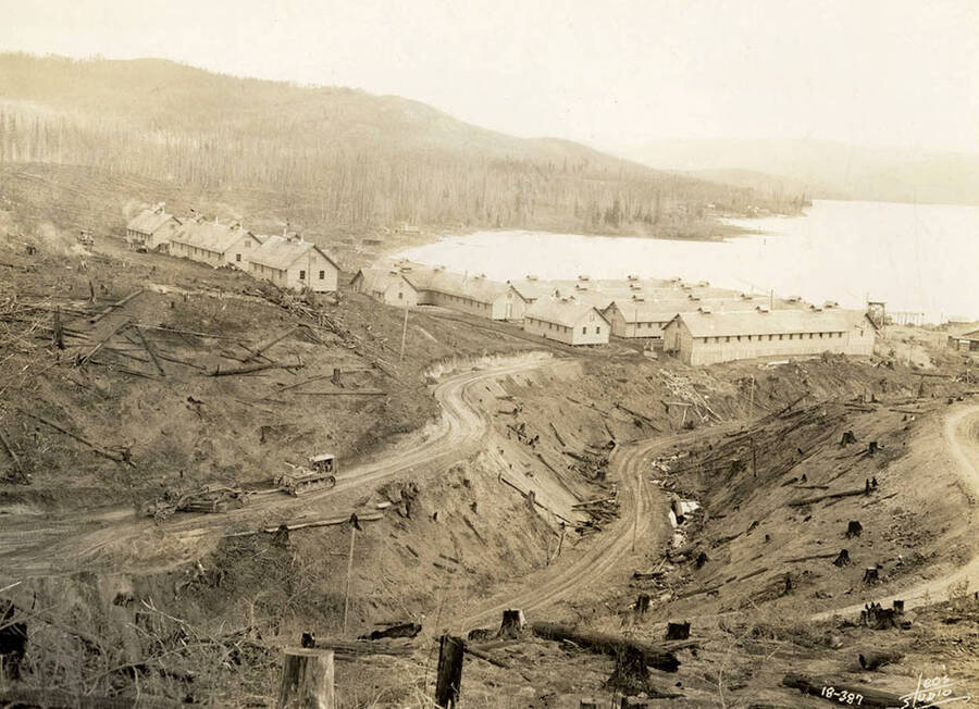 View of Kalispell Bay and the CCC Camp stationed there, F-142. Heavy equipment can be seen building a road in the foreground. Writing on the photo reads: 'Leo's Studio'. Back of photo reads: 'Kalispell Bay'