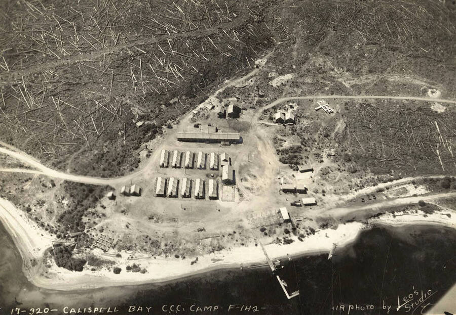 Aerial view of Kalispell Bay CCC Camp, F-142. Note the geoglyph on the right hand side of the camp it reads: 'F-142'. Writing on the photo reads: 'Calispell Bay CCC Camp F-142 Company 594. Air photo by Leo's Studio'. The 5 and 9 of 'Co. 594' has been blacked out'.