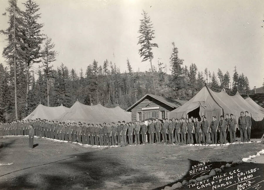 CCC men standing at attention for retreat of the flag in front of the tent barracks and an administration building at Twentymile CCC Camp, F-124. Writing on the photo reads: 'Retreat Twenty Mile CCC Camp F-124 Company 1285 Naples, Idaho Leo's Studio'. Back of photo reads: 'Kanisku'.