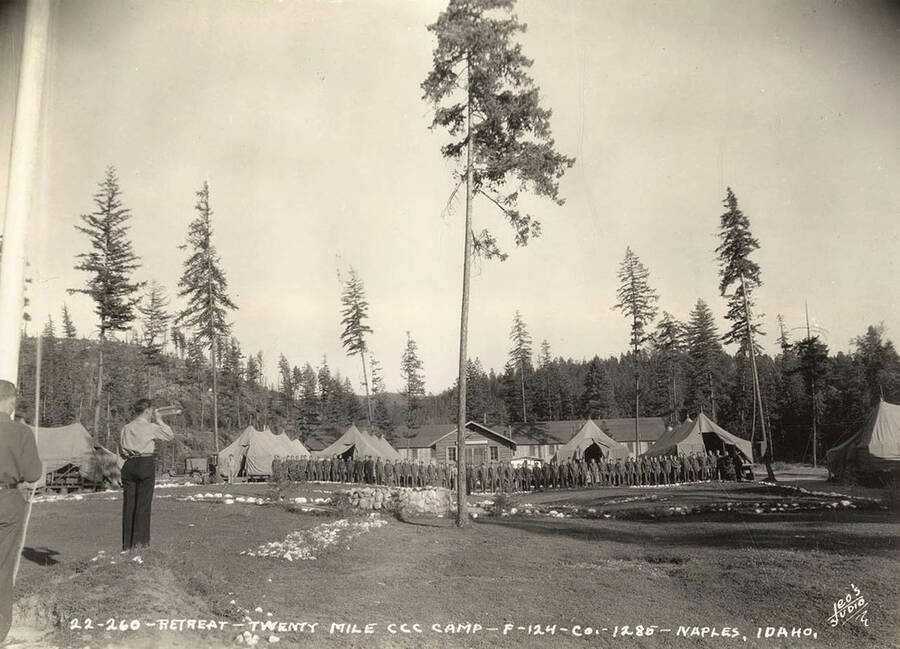 CCC men standing at attention for retreat of the flag at sundown at Twentymile CCC Camp, F-124. Note the bugler (likely playing taps) and the fountain in the center of the field. The rocks laid out likely are a geoglyph reading 'F-124'. Writing on the photo reads: 'Retreat Twenty Mile CCC Camp F-124 Company 1285 Naples, Idaho Leo's Studio'. Back of photo reads: 'Naples, Idaho'.