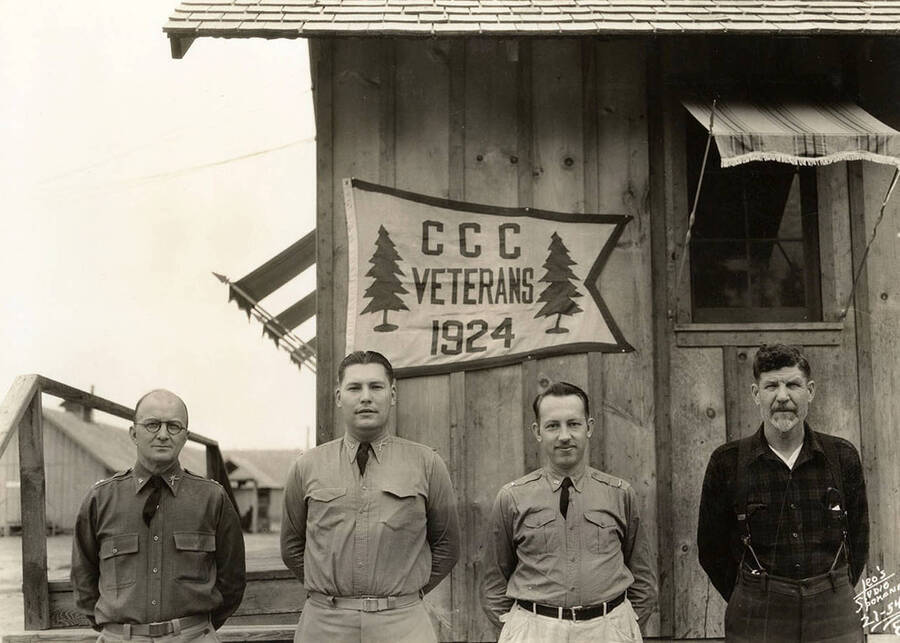 Group photo of four CCC men standing outside a building with a banner reading: 'CCC Veterans 1924'. At the Blow Down 2 CCC Camp, F-159. Back of photo reads: 'Blow Down Number 2'.