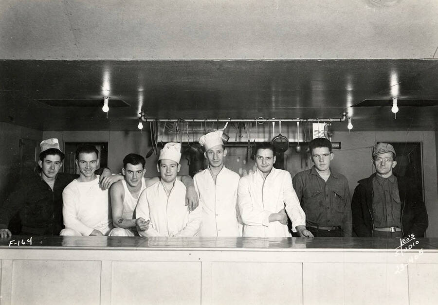 Eight CCC cooks posing in the kitchen at the Four Corners CCC Camp, F-164. Writing on the photo reads: 'F-164 Leo's Studio'. Back of photo reads: 'Four Corners Priest River Kaniksu, National Forest'.