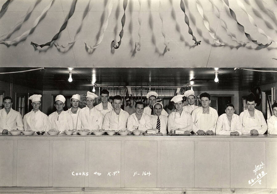 Group photo of cooks and K.P.'s (kitchen police or kitchen patrol) in the kitchen at Four Corners CCC Camp. Writing on the photo reads: 'Cooks and K.P.s F-164 Leo's Studio'. Back of photo reads: 'Four Corners Priest River Kaniksu, National Forest'.