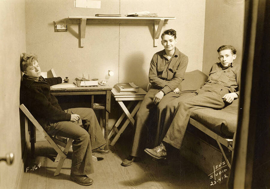 Three CCC men lounging in a dorm room at Four Corners CCC Camp, F-164. Trunk under bed reads: 'Armour'. Writing on the photo reads: 'F-164 Leo's Studio'. Back of photo reads: 'Four Corners Priest River'.