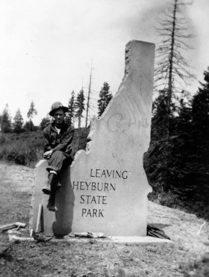 The rear view of the Heyburn State Park entrance sign. The sign reads 'Leaving Heyburn State Park'. A man is seated on the sign and there is also some graffiti reading 'C. Hill'.