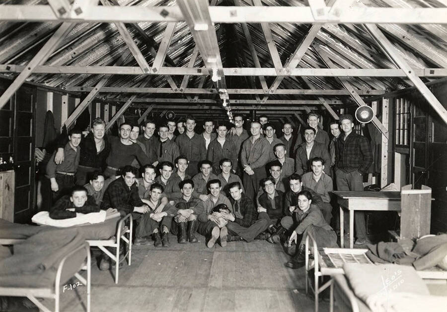 Group photo of CCC men inside a barrack at Kalispell Creek CCC Camp, F-102. Writing on the photo reads: 'F-102 Leo's Studio'. Back of photo reads: 'Kaniksu'.