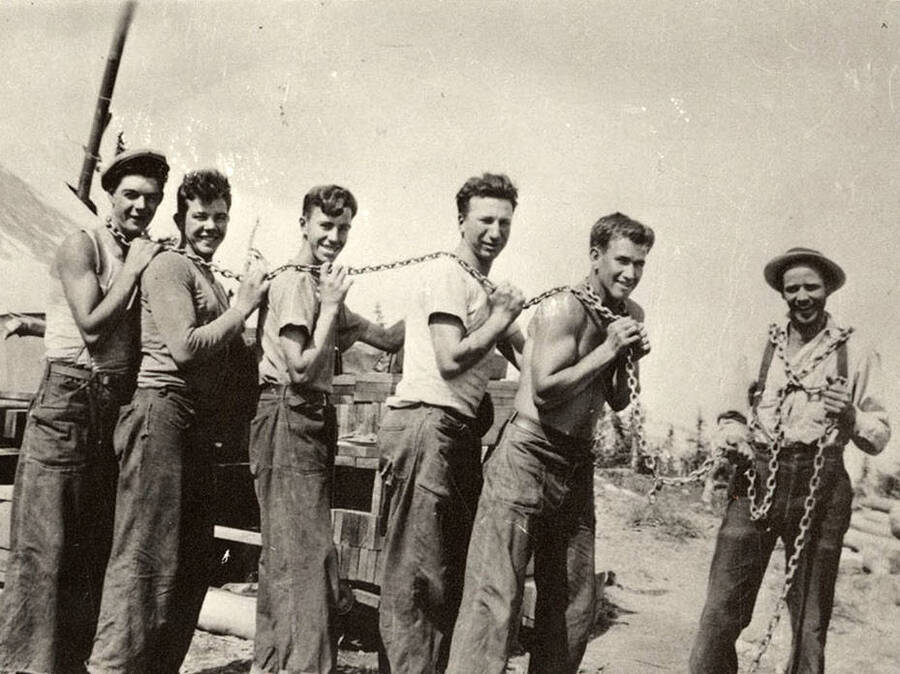 Group photo of a CCC construction crew 'The Chain Gang' posing for a photo. The man holding the chain on the right of the photo is presumably the foreman 'Jim [Parse]'. Back of the photo reads: ''The Chain Gang' Jim [Parse] Carpenter Foreman and crew of CCC's building Fire Lookout tower on Jeru Peak. 1936. Leonard Lynch photo. Chuck Peterson copy.'