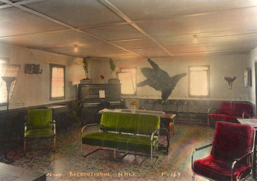Interior view of the Recreation Hall with a piano and chairs at Four Corners CCC Camp, F-164.  Note bearskin mounted on the wall. Photo has been hand painted. Writing on the photo reads: 'Recreational Hall F-164 Leo's Studio'. Back of photo reads: 'Four Corners Priest River Kaniksu National Forest'.