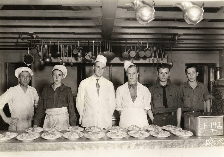 Group photo of 6 CCC men in the kitchen at Kalispell Bay CCC Camp, F-142. Writing on the photo reads F-142 Leo's Studio Spokane'. Back of the photo reads: 'Kalispell Bay'.