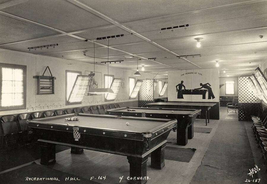 Interior view of the Recreation Hall with two pool tables and a ping pong table at Four Corners CCC Camp, F-164. Writing on the wall reads 'The reward of toil'. Note the score keeping wires hanging above the pool tables. Writing on the photo reads: 'Recreational Hall F-164 4 Corners Leo's Studio'. Back of photo reads: 'Four Corners Priest River'.