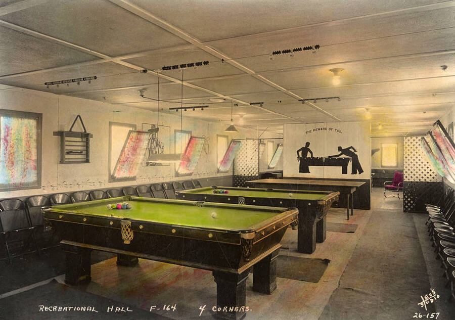 Interior view of the Recreation Hall with two pool tables and a ping pong table at Four Corners CCC Camp, F-164. Writing on the wall reads 'The reward of toil'. Note the score keeping wires hanging above the pool tables. Photo is hand painted. Writing on the photo reads: 'Recreational Hall F-164 4 Corners Leo's Studio'. Back of photo reads: 'Four Corners Priest River'.