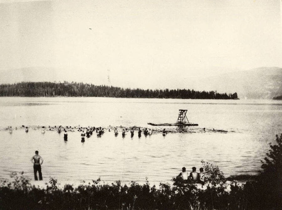 View of CCC men swimming in Kalispell Bay. There is a life guard standing watch and a row of three men sitting on the shore in the foreground. Back of photo reads: 'CCC Swimming lessons at Kalispell Bay, Headquarters Camp F-142, 1935. Boys were brought to Priest Lake from other camps for swimming lessons. Les [Tam] photo. Chuck Peterson copy.