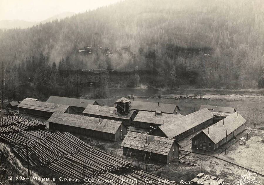 View of Marble Creek CCC Camp, F-117, under construction. Also shown is Marble Creek and piles of logs next to the camp buildings. Writing on the photo reads: 'Marble Creek CCC Camp F-117 Company 240 October 20, 1934 Leo's Studio Spokane'.