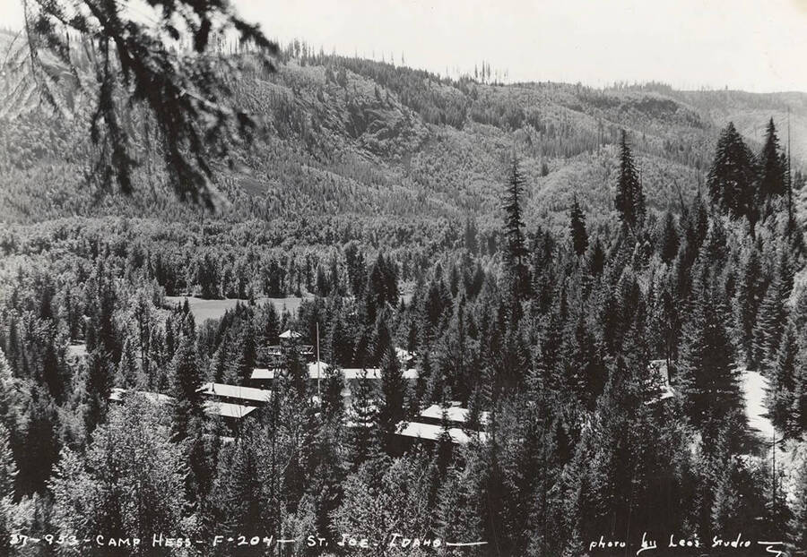A view of CCC Camp Hess, F-204 through the trees and the surrounding forest. Note the water tower. Writing on the photo reads: 'Camp Hess F-204 St. Joe, Idaho. Photo by Leo's Studio'. Back of photo reads: Camp Hess St. Joe'.