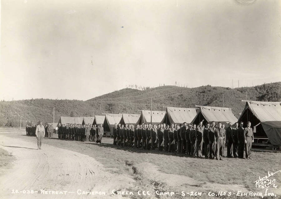 View of CCC men standing at attention during retreat of the flag at Cameron Creek CCC Camp, S-264. Writing on the photo reads: 'Retreat Cameron Creek CCC Camp S-264 Company 1653 Elk River, Idaho Photo by Leo's Studio'.