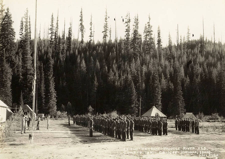 View of CCC men standing at attention for retreat of the flag at Palouse River CCC Camp, F-185 (or P-185). Writing on the photo reads 'Retreat Palouse River CCC Camp F-185 Company 1655 Harvard, Idaho Leo's Studio'.