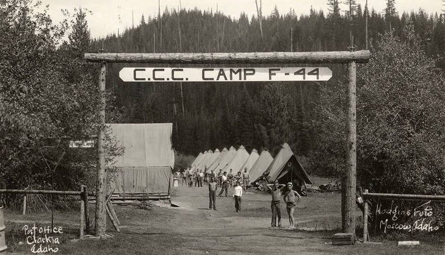 View of CCC men posing under a sign for Merry Creek CCC Camp F-44 in front of the rest of the camp. Large sign reads: 'CCC Camp F-44' Sign to the left reads: 'Fire alarm'. Writing on the photo reads: 'Post Office Clarkia, Idaho Hodgins Foto Moscow, Idaho'. Back of photo reads: 'Merry Creek 1933'.