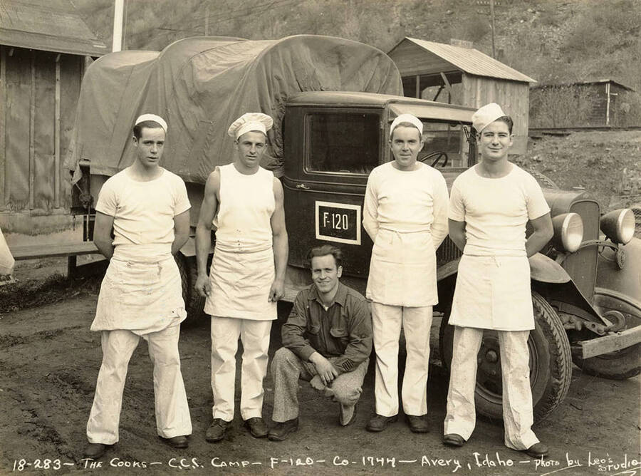 Group photo of cooks posed in front of a truck at CCC Camp F-120, near Avery, Idaho. Sign on truck reads: 'F-120'. Writing on the photo reads: 'The cooks CCC Camp F-120 Company 1744 Avery, Idaho photo by Leo's Studio'. Back of photo reads: 'Avery'. Stamp on back of photo reads: 'Nov 20 1934 SHIP'D'.