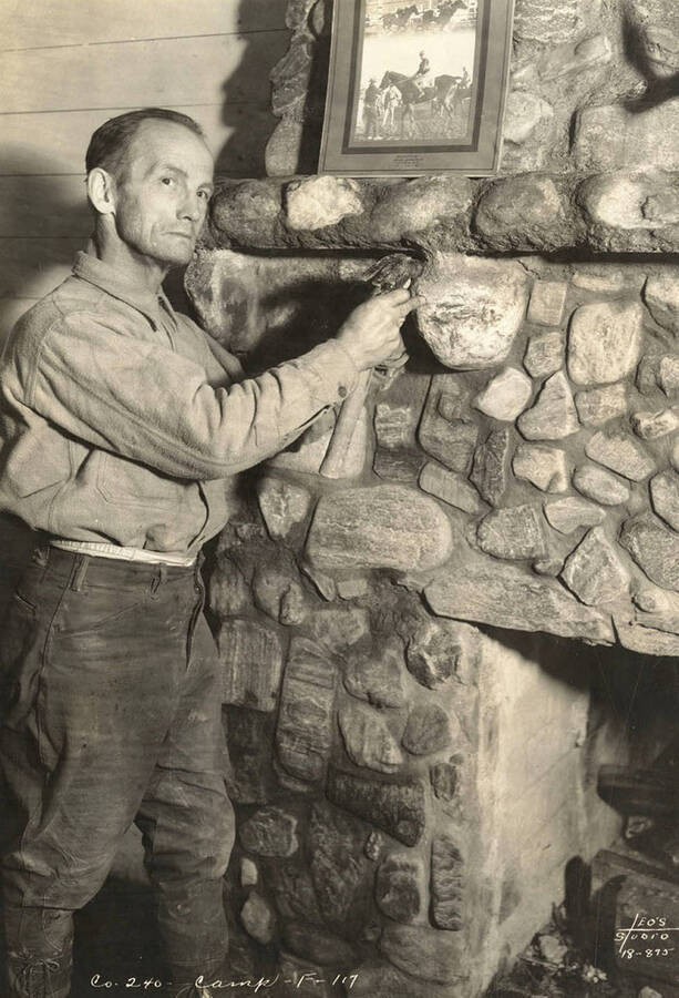 CCC Man from Marble Creek CCC Camp, F-117, posed at a stone fireplace with a hammer in his hand. Writing on the photo reads: ' Company 240 Camp F-117 Leo's Studio'. Back of photo reads 'Marble Creek'.