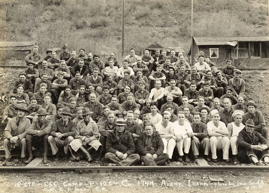 Group photo of CCC men in Company 1744 sitting on the railroad tracks at CCC Camp F-120 near Avery, Idaho. Writing on the photo reads: 'CCC Camp F-120 Company 1744 Avery, Idaho photo by Leo's Studio 10/19/34 Captain C.F. Jago C.O.' Back of photo reads: 'Avery'.