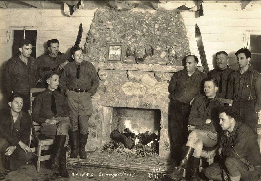 Group photo of CCC men arranged around a stone fireplace at the Marble Creek CCC Camp, F-117. Plaque above the fireplace reads: 'Company 245 [or possibly 240] CCC'. Writing on the photo reads: 'Company 240 Camp F-117 Leo's Studio'.