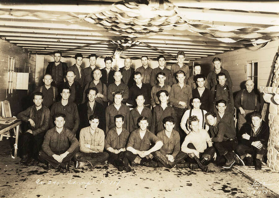 Group photo of CCC men posed in a building at Marble Creek CCC Camp, F-117. Writing on the photo reads: 'Company 240 Camp F-117 Leo's Studio'.