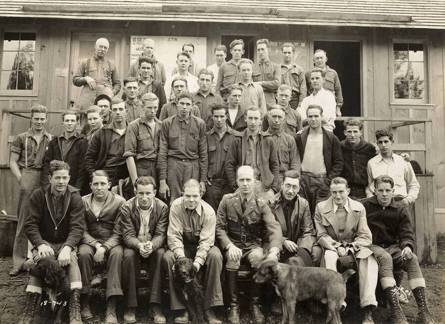 Group of CCC men posing for a photo with three dogs outside of a building at CCC Camp Heyburn, SP-1, Company 1995. Franklyn W. Bovey, educational, bottom row second from the end on the left. Note the bulletin board on the side of the building behind the men. One of the bulletins reads: 'One Night Only Al Freeman's Floor Show'. Writing on photo reads: 'Leo's Studio'. Back of photo reads: 'Pettee'.