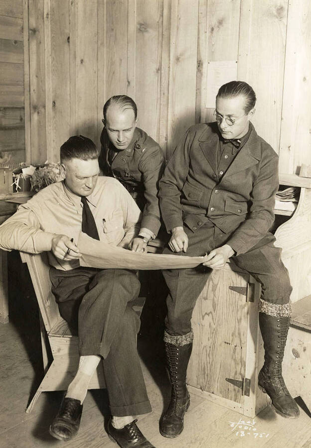 Group photo of three officers observing a document or map in an office at CCC Camp Heyburn, SP-1. Writing on the photo reads: 'Leo's Studio' Back of photo reads: 'Heyburn'.