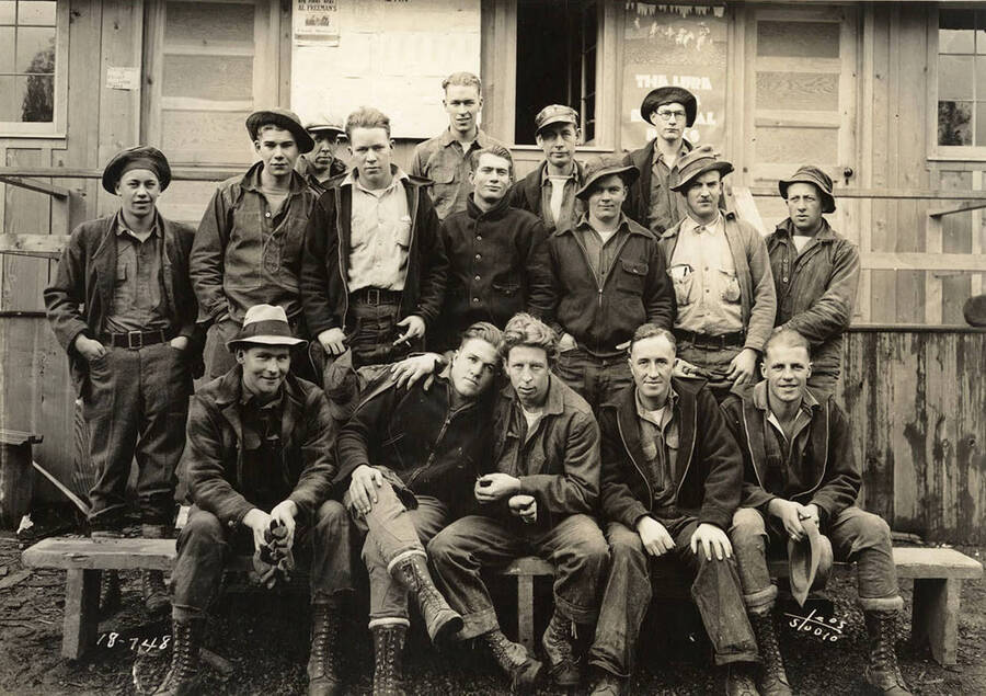 Group of CCC men posing for a photo outside of a building at CCC Camp Heyburn, SP-1, Company 1995. Bottom row, fourth from left is Ray Wozny. Note the bulletin board on the side of the building behind the men. One of the bulletins reads: 'One Night Only Al Freeman's Floor Show'. Poster reads: 'The Lure of the National Parks'. Writing on the photo reads: 'Leo's Studio'. Back of photo reads: 'Heyburn'.