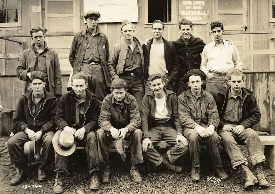 Group of CCC men posing for a photo outside one of the buildings at CCC Camp Heyburn, SP-1, Company 1995. Note the bulletin board on the side of the building behind the men. Poster reads: 'The Lure of the National Parks'.  Back of photo reads: 'Heyburn'.