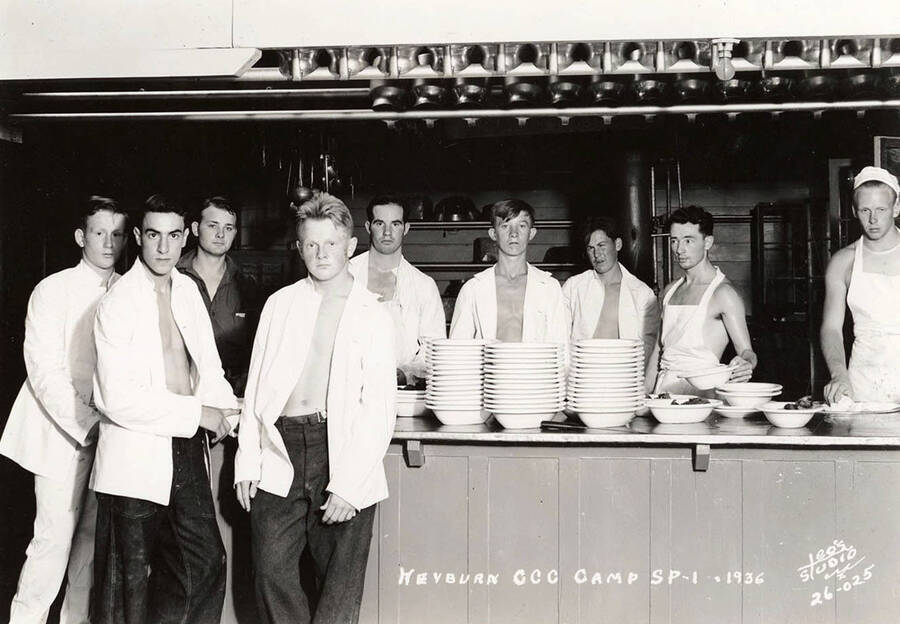 Group photo of CCC Camp Heyburn cooks and K.P.s posing in the kitchen, SP-1, Company 1995. From left to right: Front Row: Paul Lahaih, with his right arm crossed over his body, Milsen Kellner. Back Row: Unidentified man, Gerald Stewart, Joe Deroshia, unidentified man, unidentified man, Harry Nelson, and Morton Rolf. Writing on the photo reads: 'Heyburn CCC Camp SP-1 1936 Leo's Studio'. Back of photo reads: 'Heyburn'.