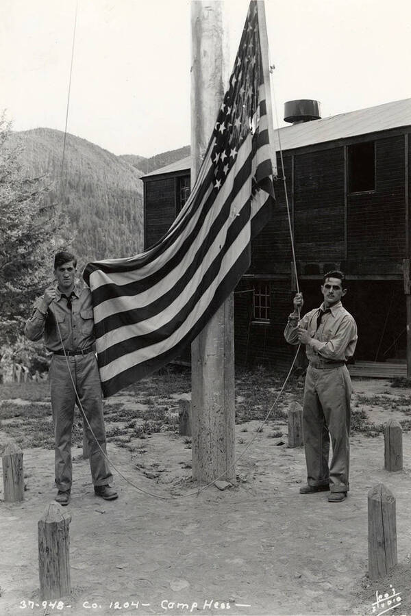 Two CCC men posing with the American flag at CCC Camp Hess, F-204, Company 1204. Writing on the photo reads: 'Company 1204 Camp Hess Leo's Studio'. Back of photo reads 'Camp Hess St. Joe'.