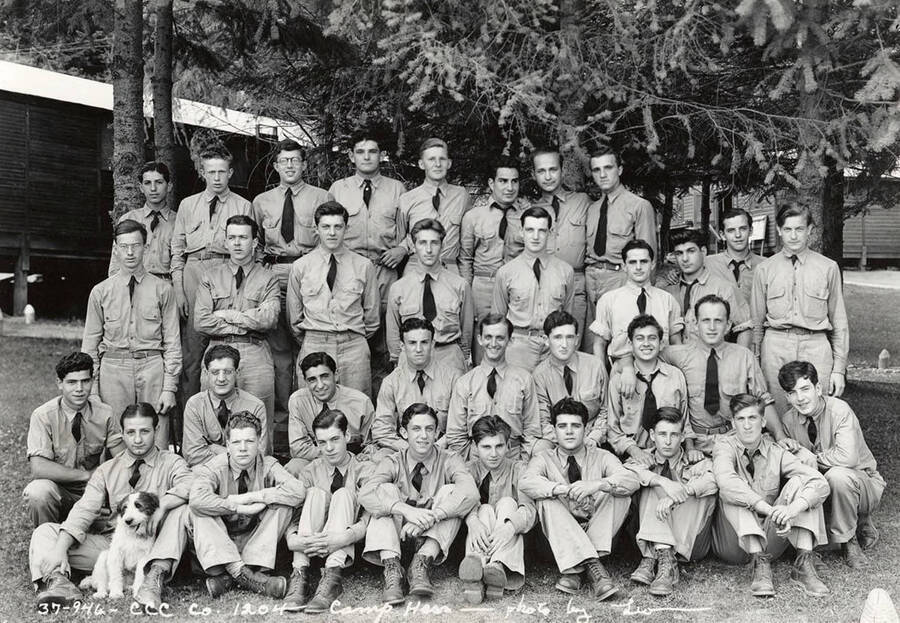 Group of uniformed CCC men posing for a photo with a dog at CCC Camp Hess, F-204, Company 1204. Writing on the photo reads: 'CCC Company 1204 Camp Hess photo by Leo'. Back of photo reads: 'Camp Hess St. Joe'.