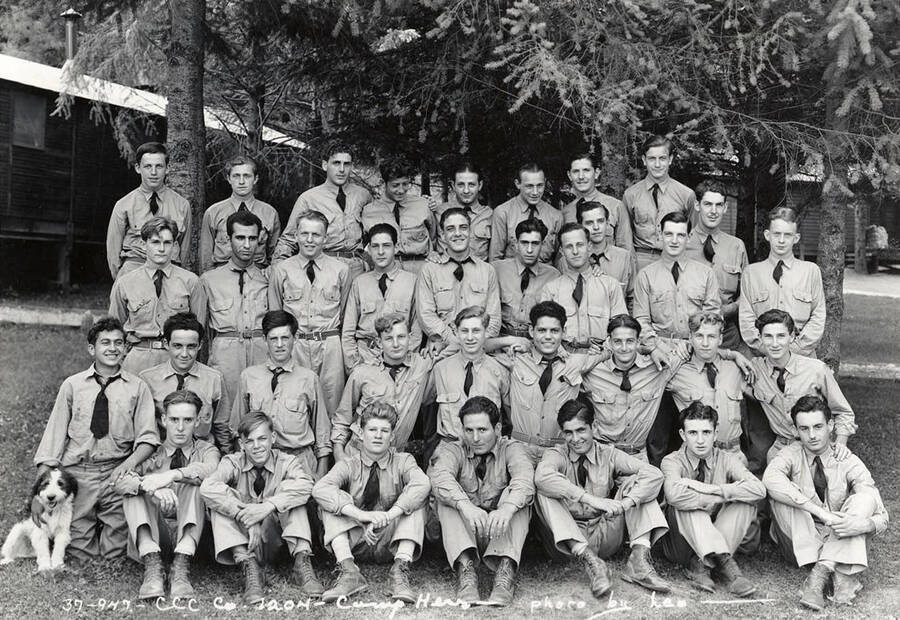 Group of uniformed CCC men posing for a photo with a dog at CCC Camp Hess, F-204, Company 1204. Writing on the photo reads: 'CCC Company 1204 Camp Hess photo by Leo'. Back of photo reads: 'Camp Hess St. Joe'.