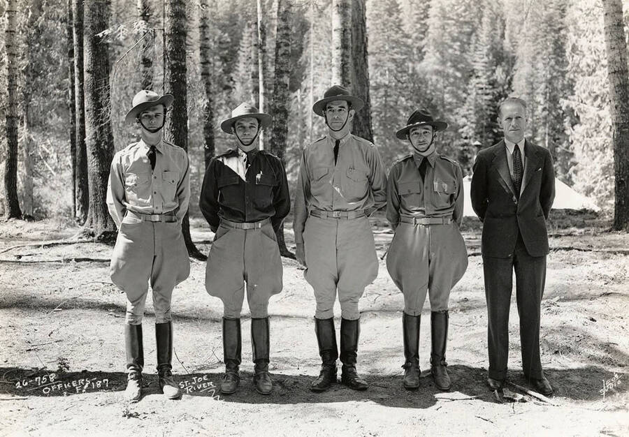 Group photo of four CCC officers standing in a row in the woods with a man in a suit near St. Joe River CCC Camp, F-187, Company 1239. Writing on the photo reads: 'Officers F-187 St. Joe River Leo's Studio'. Back of photo reads: 'St. Joe River F-187'.
