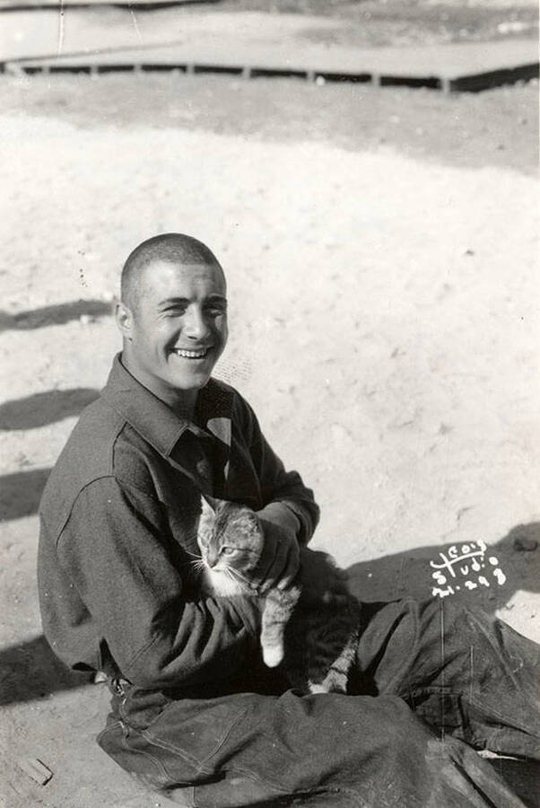 CCC man posed with a cat at Merry Creek CCC Camp, F-140. Writing on the photo reads: 'Leo's Studio'. Back of photo reads: 'St. Joe'.