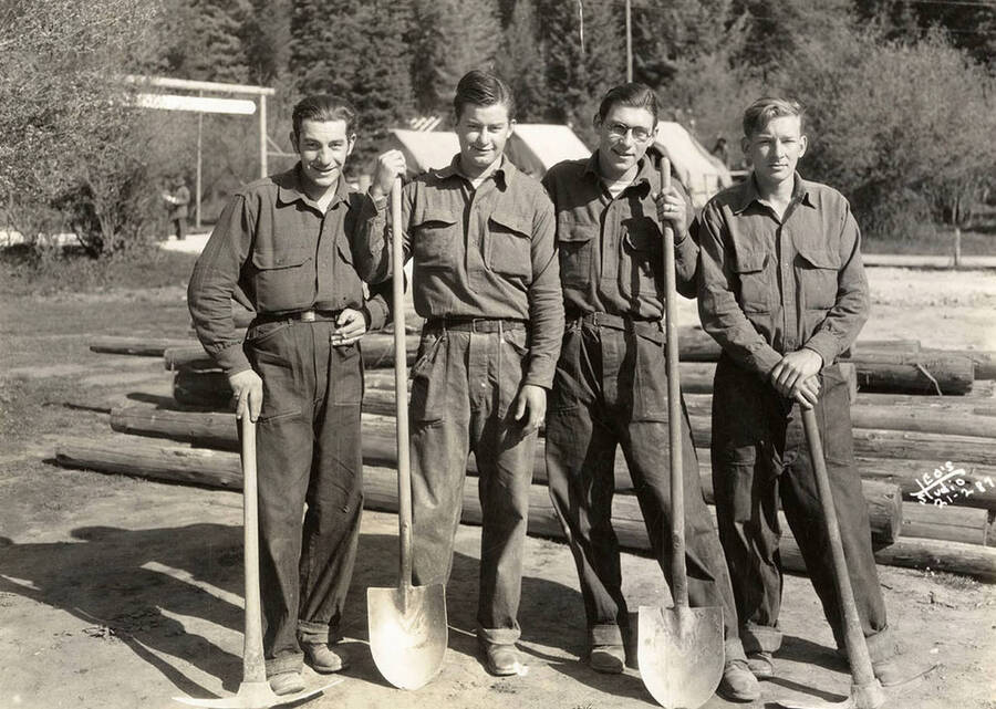 Group photo of four CCC men posed with shovels and hand picks at Merry Creek CCC Camp, F-140, in front of a log pile with tents in the background. Writing on photo reads: 'Leo's Studio'. Back of photo reads: 'St. Joe Merry Creek'.