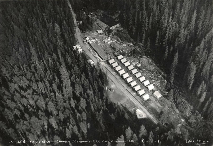 Aerial view of Badger Meadows CCC Camp, F-158 Company 557. Roof of building reads: 'F-158'. Next to road two words are visible, they read: 'Indiana' and 'Idaho'. At the top of the photo you can see that the creek has been dammed to form a swimming pond. Writing on the photo reads: 'Air view Badger Meadows CCC Camp F-158 Company 557 Leo's Studio'. Back of photo reads: 'Clearwater'.